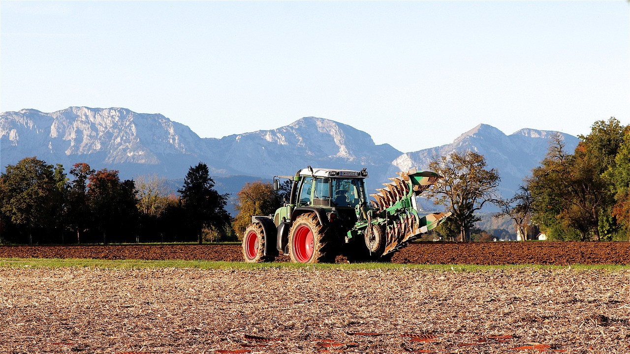 Grüner Traktor mit Pflug auf Acker vor Gebirgskulisse.